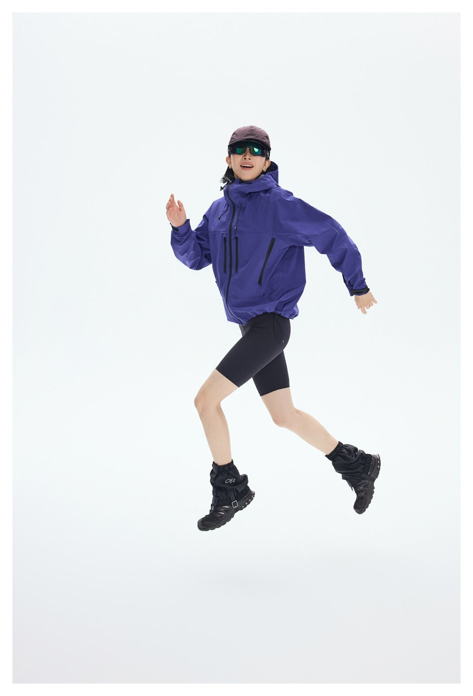 A woman in a purple hooded jacket and black shorts poses mid-stride against a plain light background.