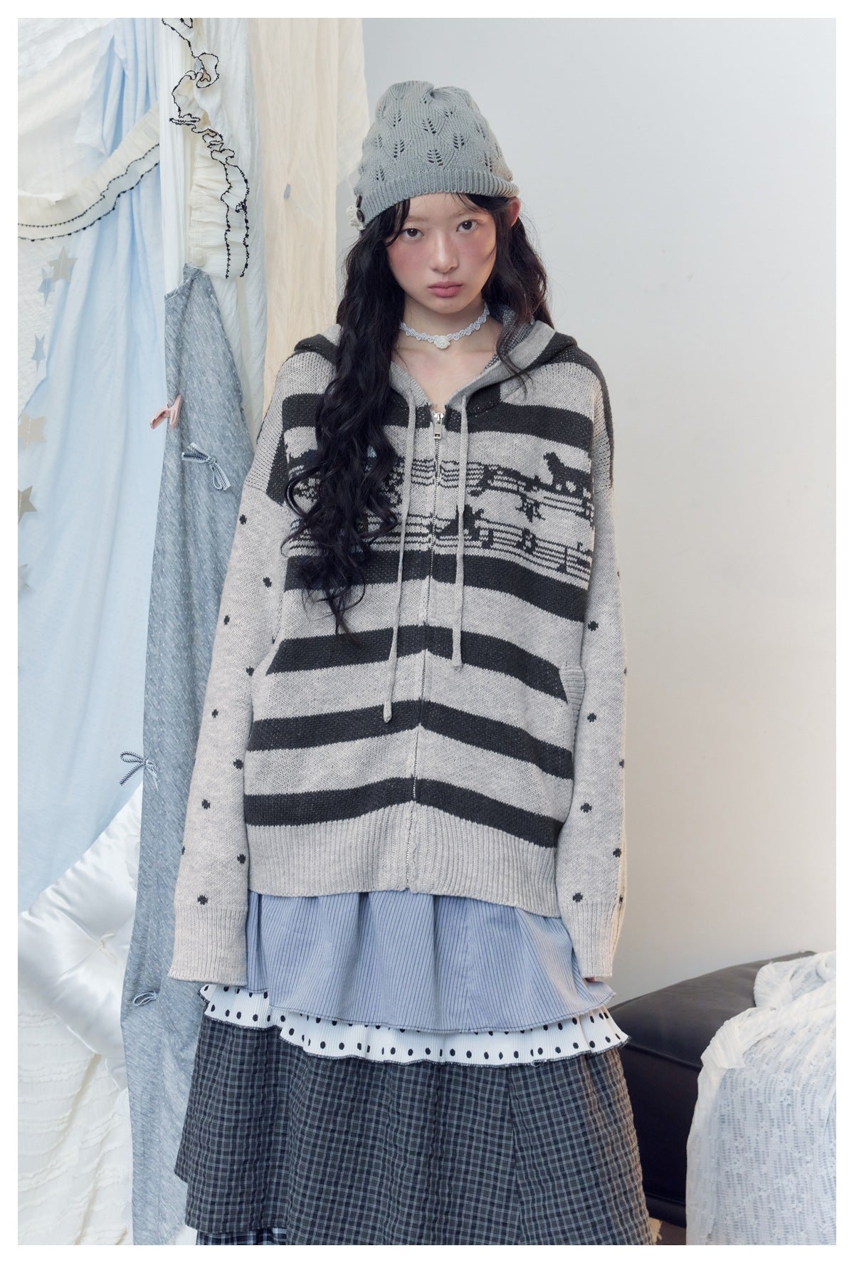 Model wearing a gray zip-up hooded sweater with black stripes, a patterned chest band and dotted sleeves over a layered