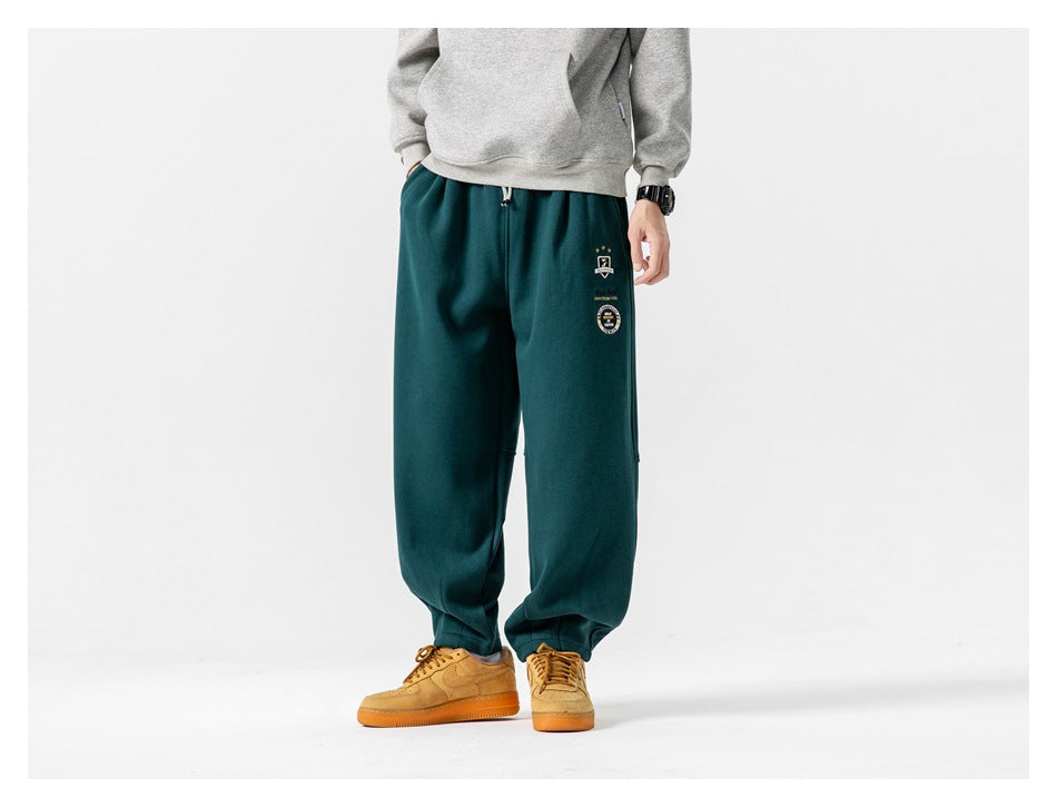 Front view of a model wearing deep teal joggers with a drawstring waist, gathered cuffs and small embroidered leg graphics.
