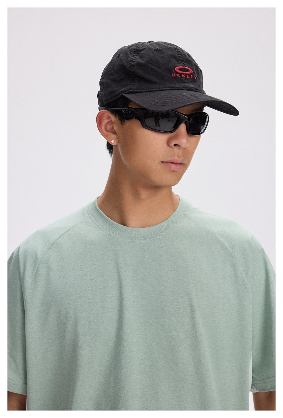 Model wearing a plain pale sage short-sleeve crewneck T-shirt, shown from the front; a black cap and sunglasses are also