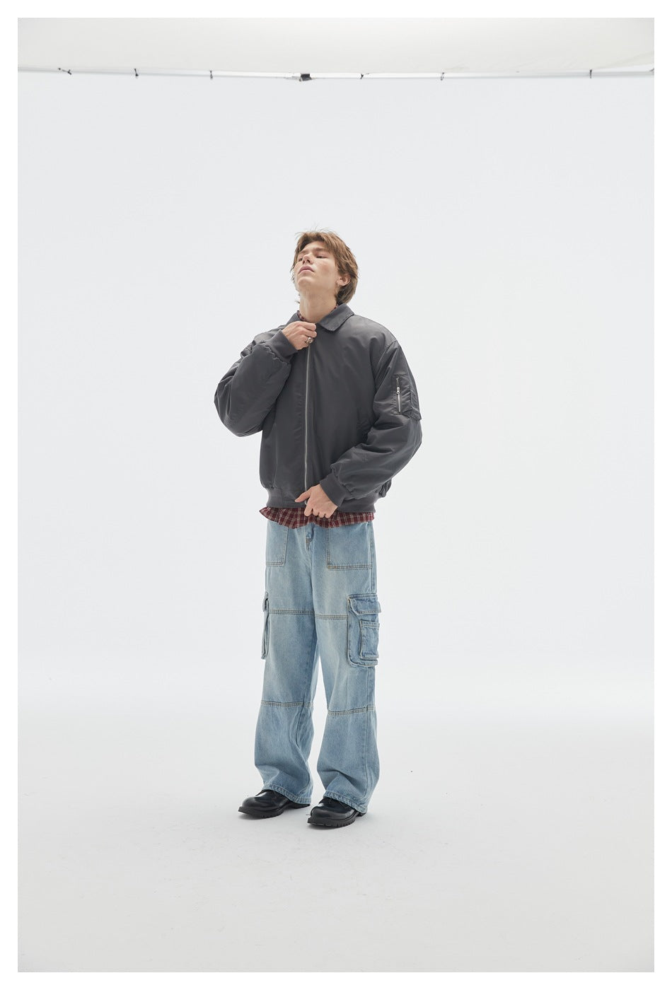 Model wearing a dark gray zip bomber jacket with a sleeve pocket over a plaid shirt and light-wash cargo jeans