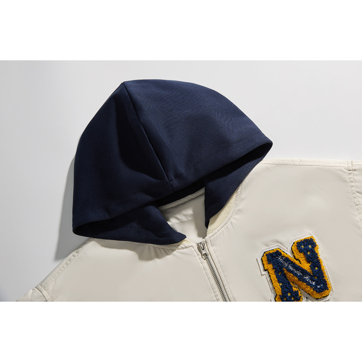 Close-up of an ivory zip-up jacket with a dark navy hood and a blue-and-gold embroidered letter patch.