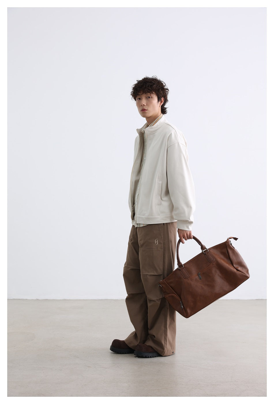 Model wearing an off-white zip jacket and brown cargo pants, carrying a large brown bag.