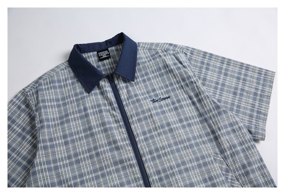 Blue-gray plaid short-sleeve zip-up shirt laid flat, showing its navy collar and small chest embroidery.