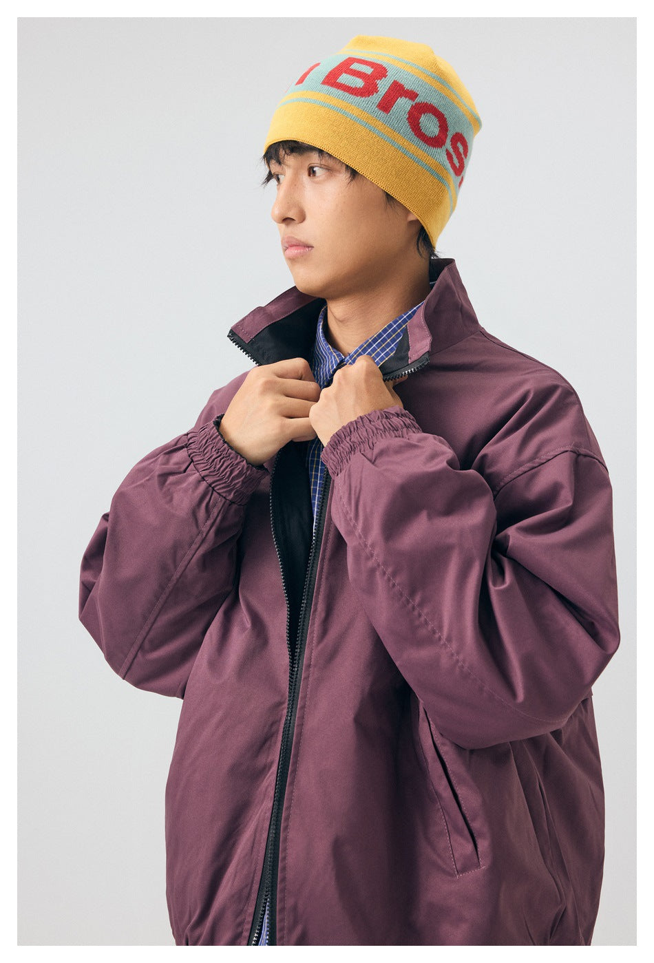 Close view of a model adjusting the collar of a plum zip jacket; a yellow graphic beanie and blue checked shirt are visible.