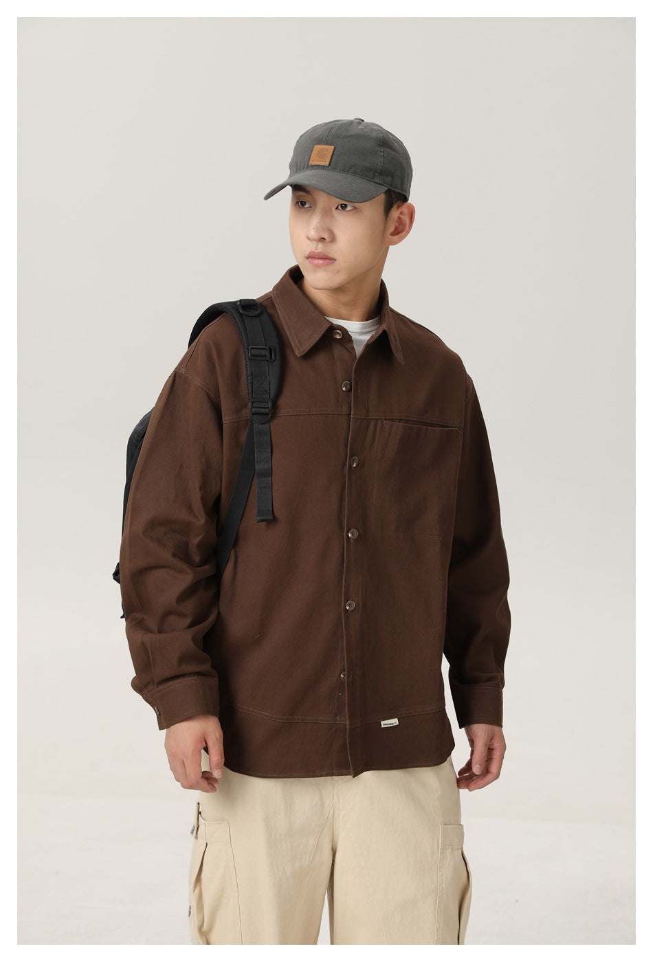 Closer front view of a model wearing a dark brown button-front shirt jacket, white tee, and dark cap.