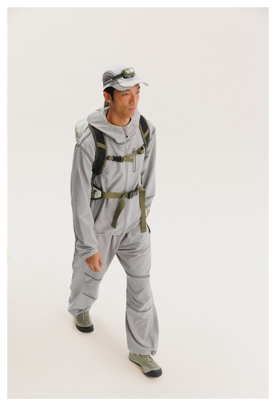 Model in a light gray hooded outfit with darker trim, an olive backpack, and a cap-mounted headlamp.
