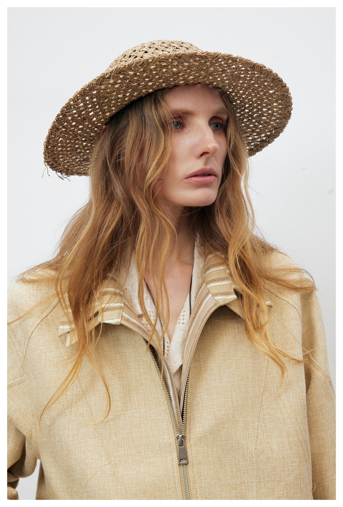Model wearing a beige zip-front jacket with a striped collar and a wide woven brown hat.