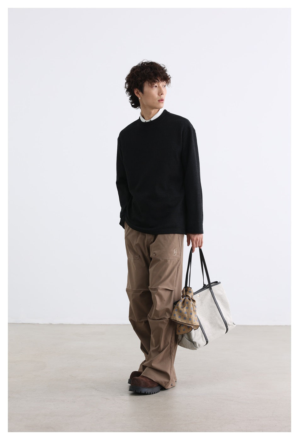 Model in a black ribbed crewneck and brown trousers, carrying a light tote bag with dark handles.