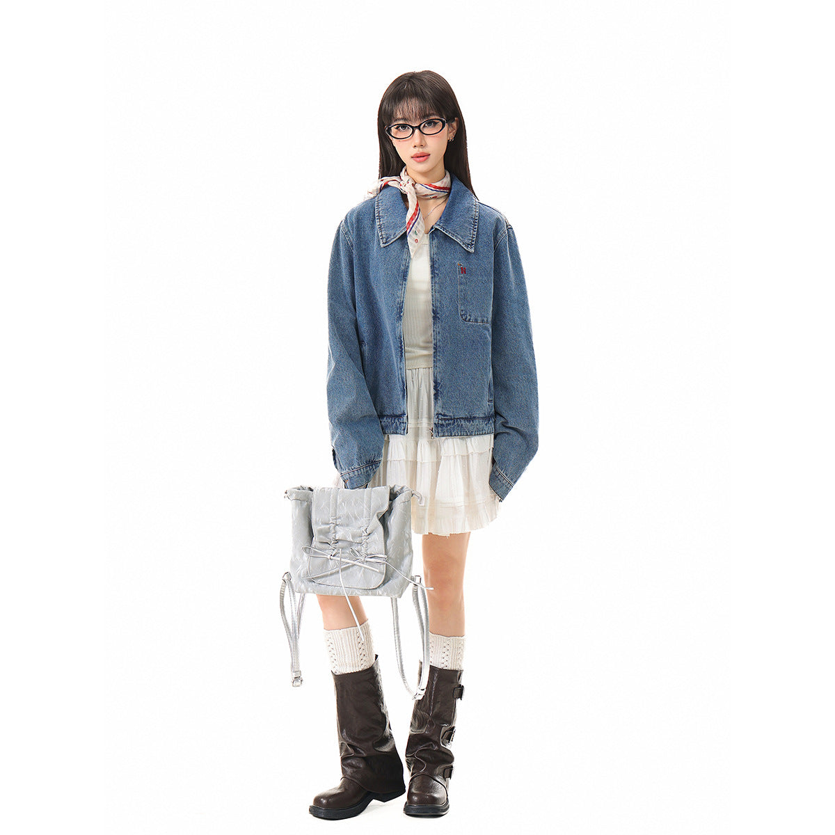 Full-length model wearing an oversized blue denim jacket over a white ruffled dress, with tall dark boots and a silver bag.