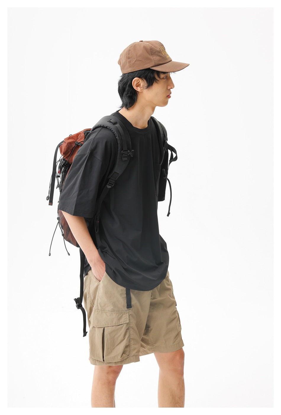 Model wearing a loose black T-shirt and tan cargo shorts with a cap and hiking backpack.