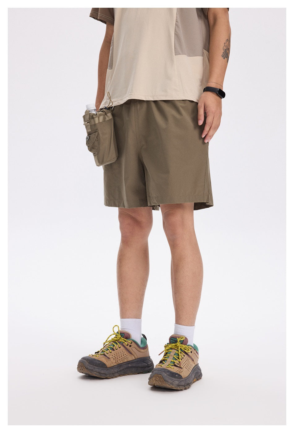 Person wearing olive green shorts with a bottle pouch at the hip, beige top and hiking shoes, cropped front view