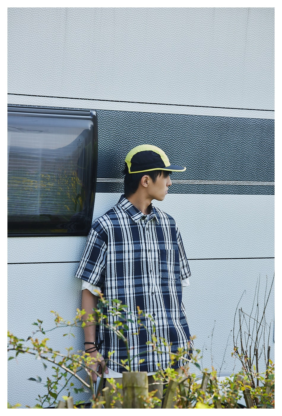 Side view of a model in an oversized navy and white plaid shirt, with a neon yellow and black cap.