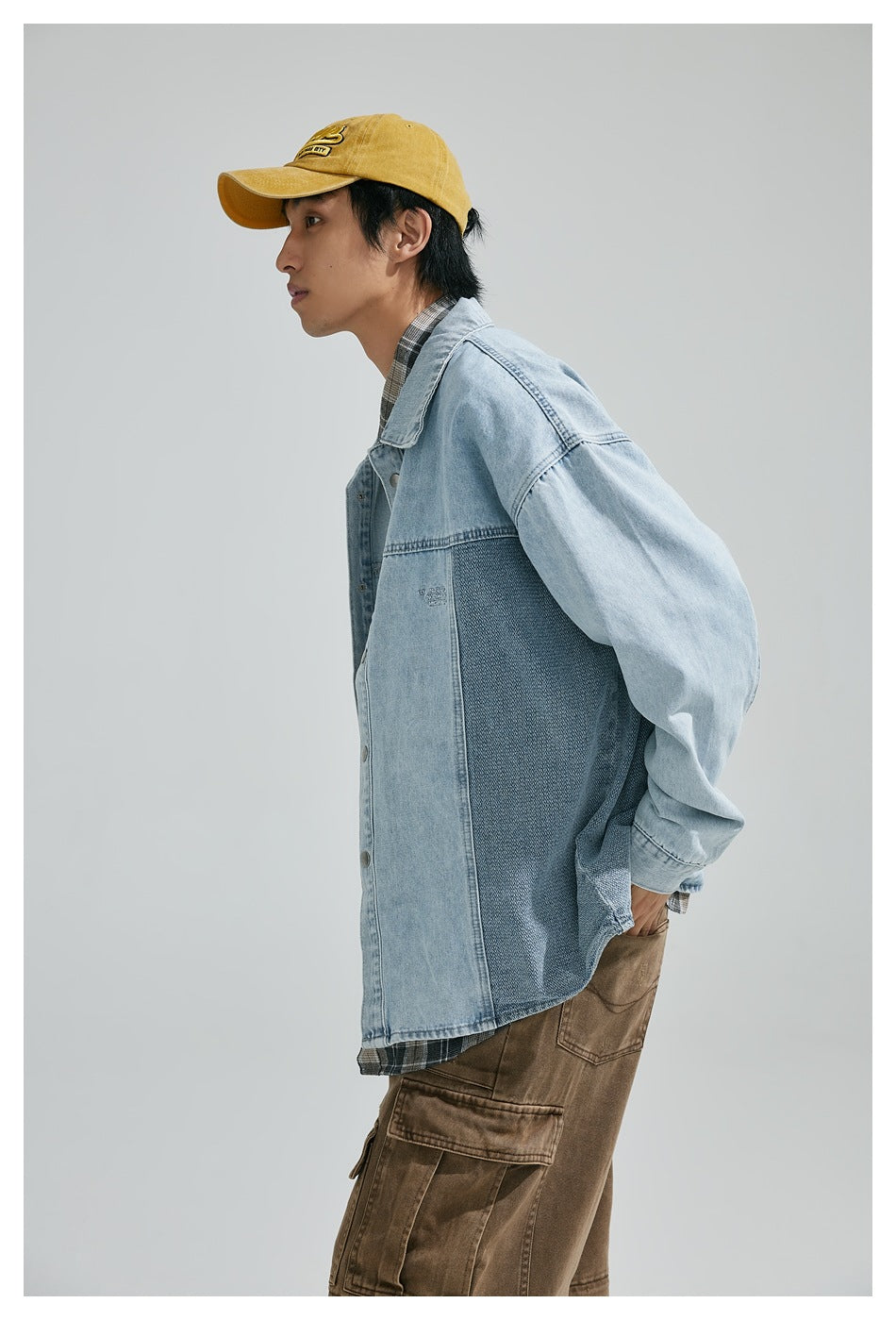 Model in side view wearing a light blue color-block denim shirt jacket, plaid shirt, brown cargo pants, and mustard cap.