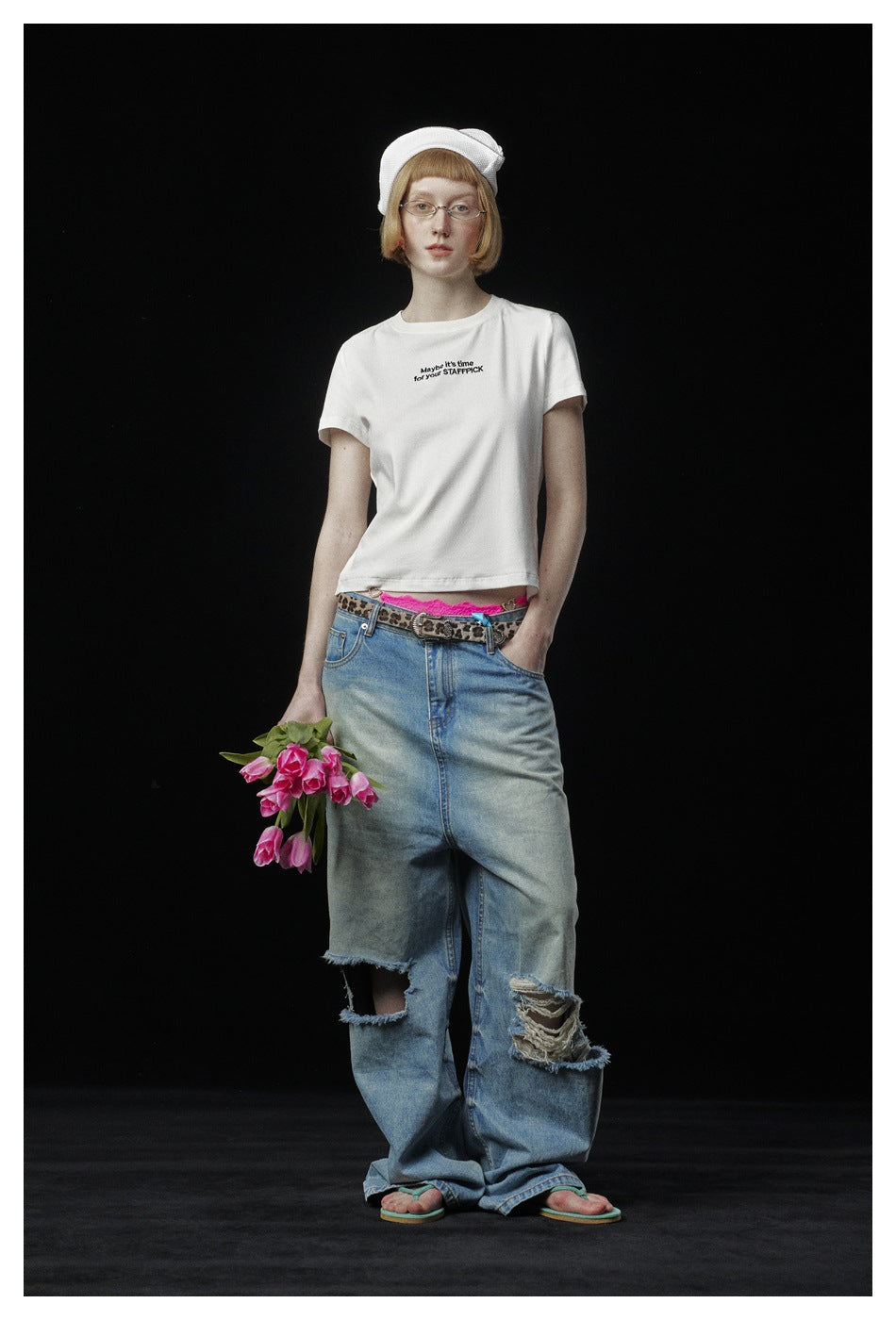 Model wears a white slogan T-shirt and distressed wide-leg jeans with a white cap, holding pink tulips against black.