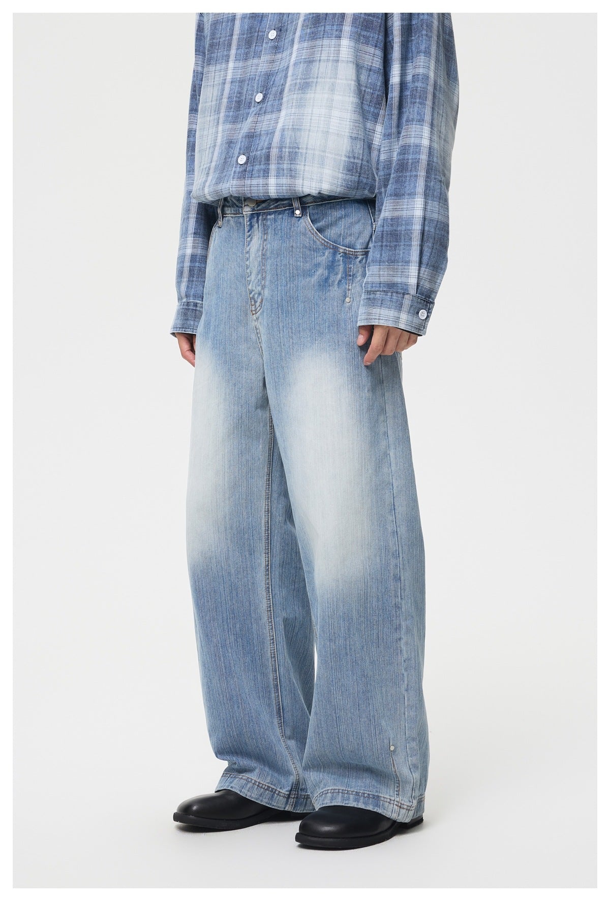 Cropped view of a person wearing light blue faded, wide-leg jeans with a blue plaid shirt and black shoes, front view.