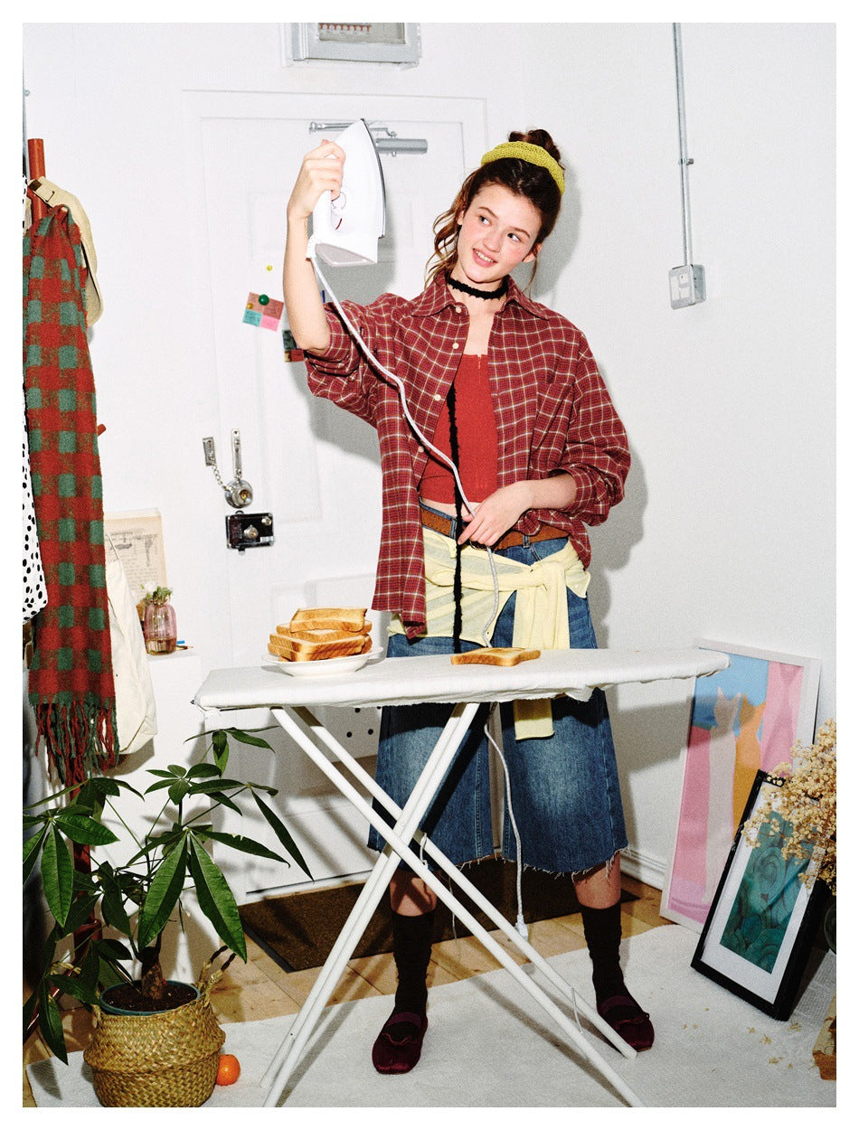 Model wearing a red plaid button-up shirt over a red top, holding an iron beside an ironing board.