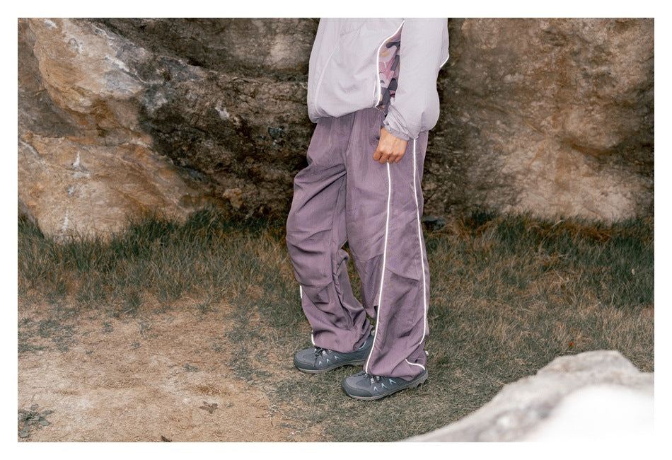 Person wearing purple wide-leg pants with white side piping, cropped from chest down, standing outdoors by rocks