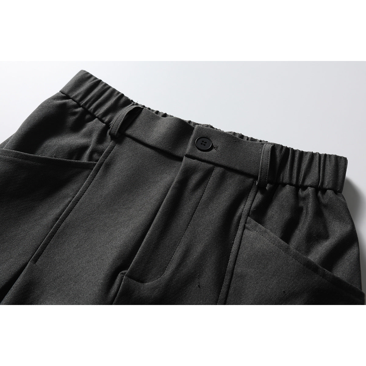Close-up of dark charcoal trousers showing an elasticated waistband, belt loops, button closure and front pocket.