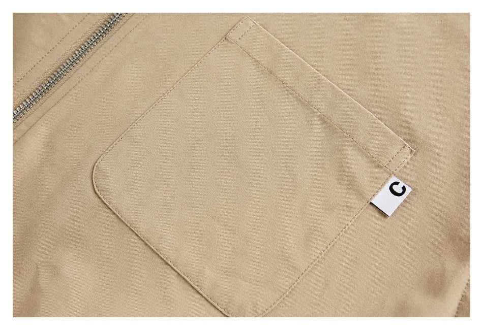 Close-up of a tan jacket's patch pocket, small white tab, and metal zipper.
