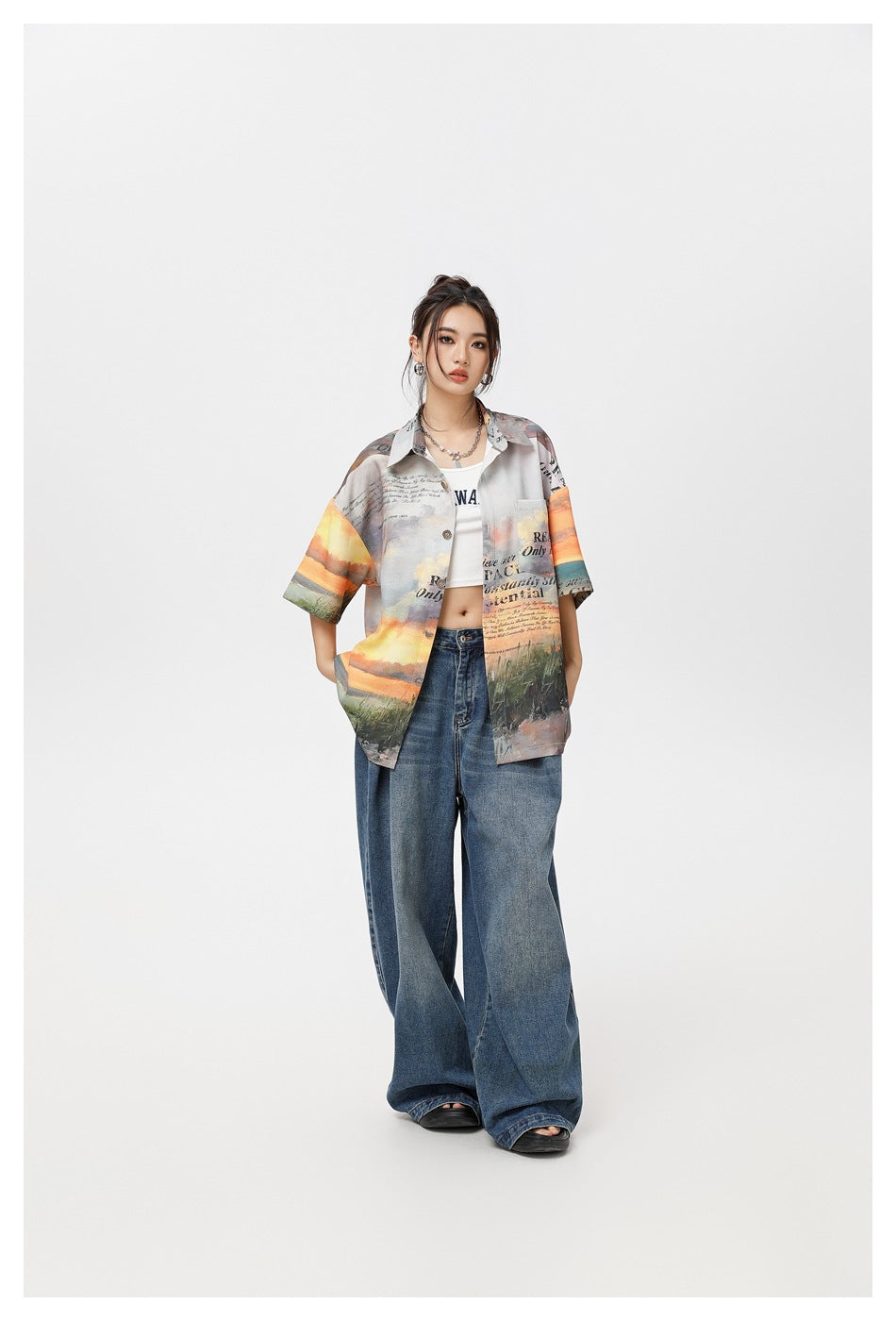 Model wearing an open sunset-print short-sleeve shirt over a white crop top with wide-leg jeans, hands in pockets