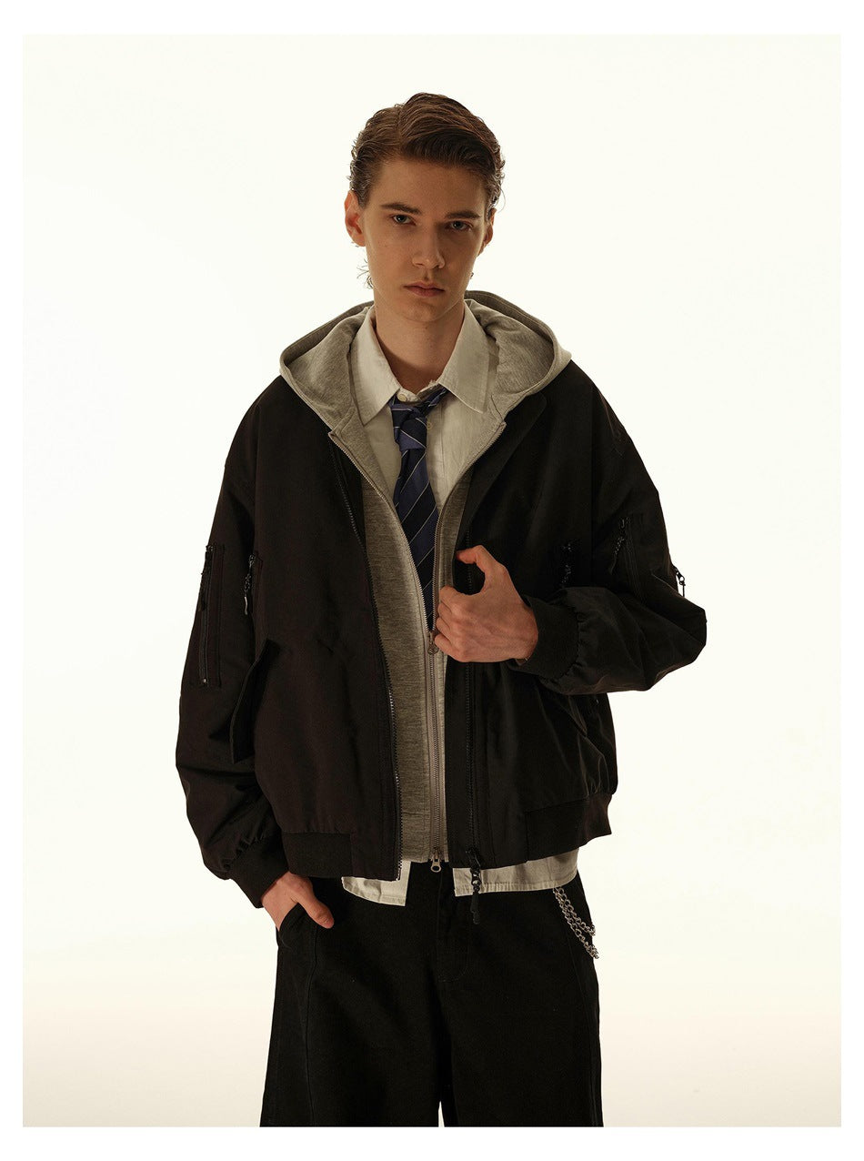 Man modeling a black bomber jacket over a gray hoodie, shown from the waist up with the jacket partly open.