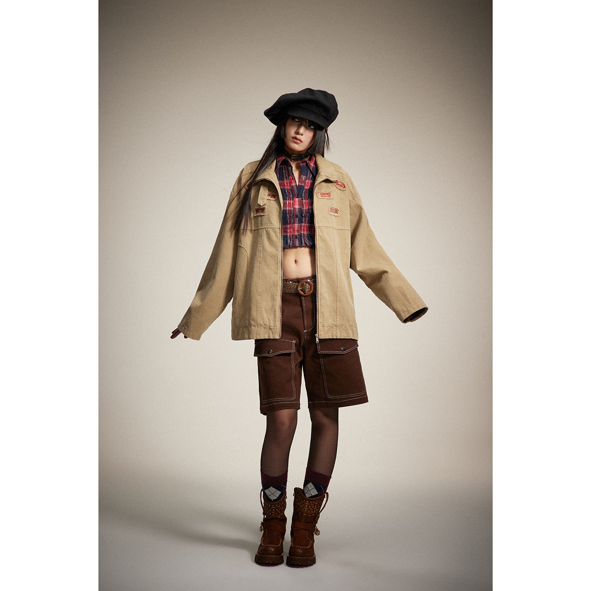 Model wearing a tan patch-decorated zip jacket over a red plaid crop top and brown cargo shorts.