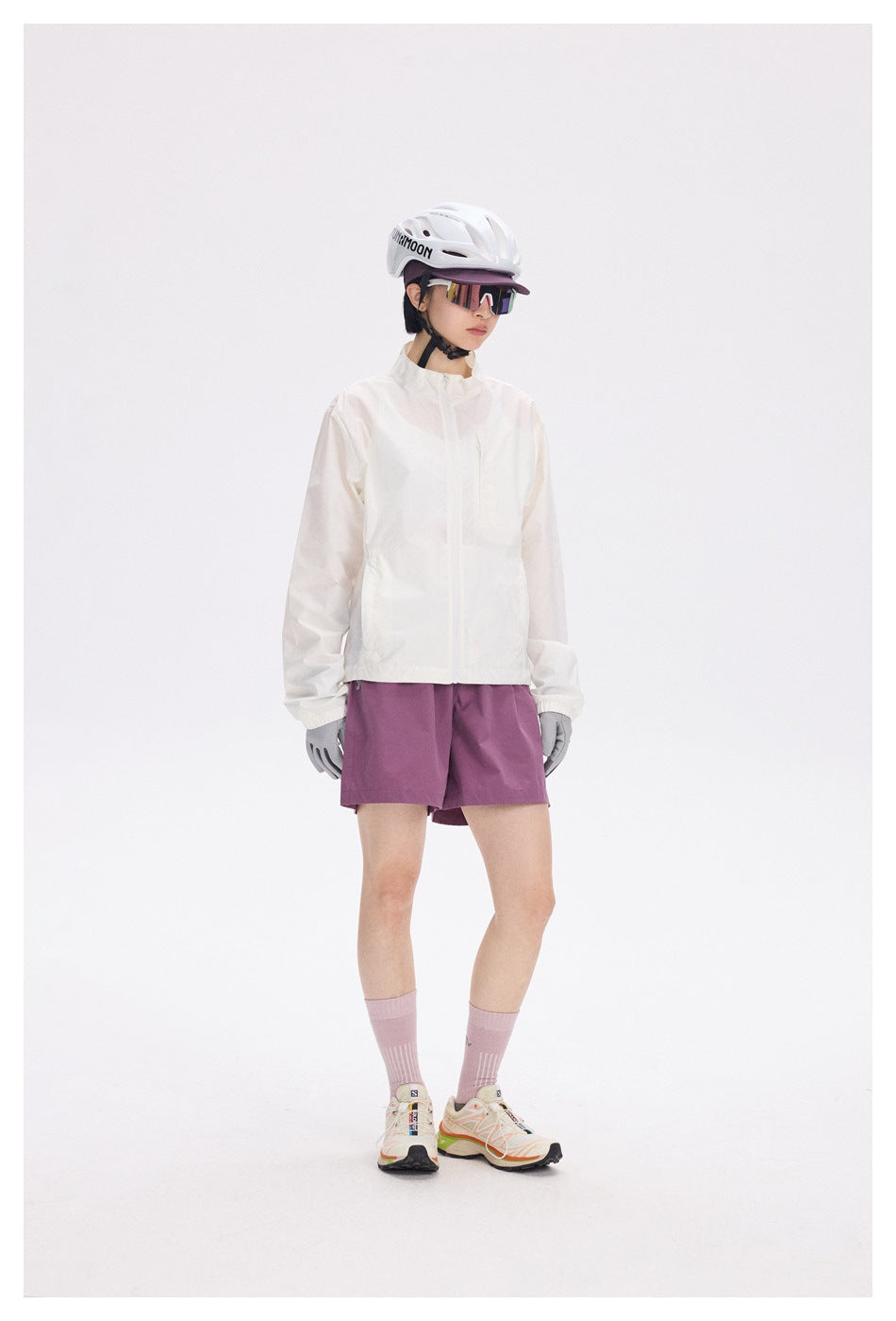 Model wearing a zipped white stand-collar jacket with purple shorts, helmet, gloves and sneakers, angled view