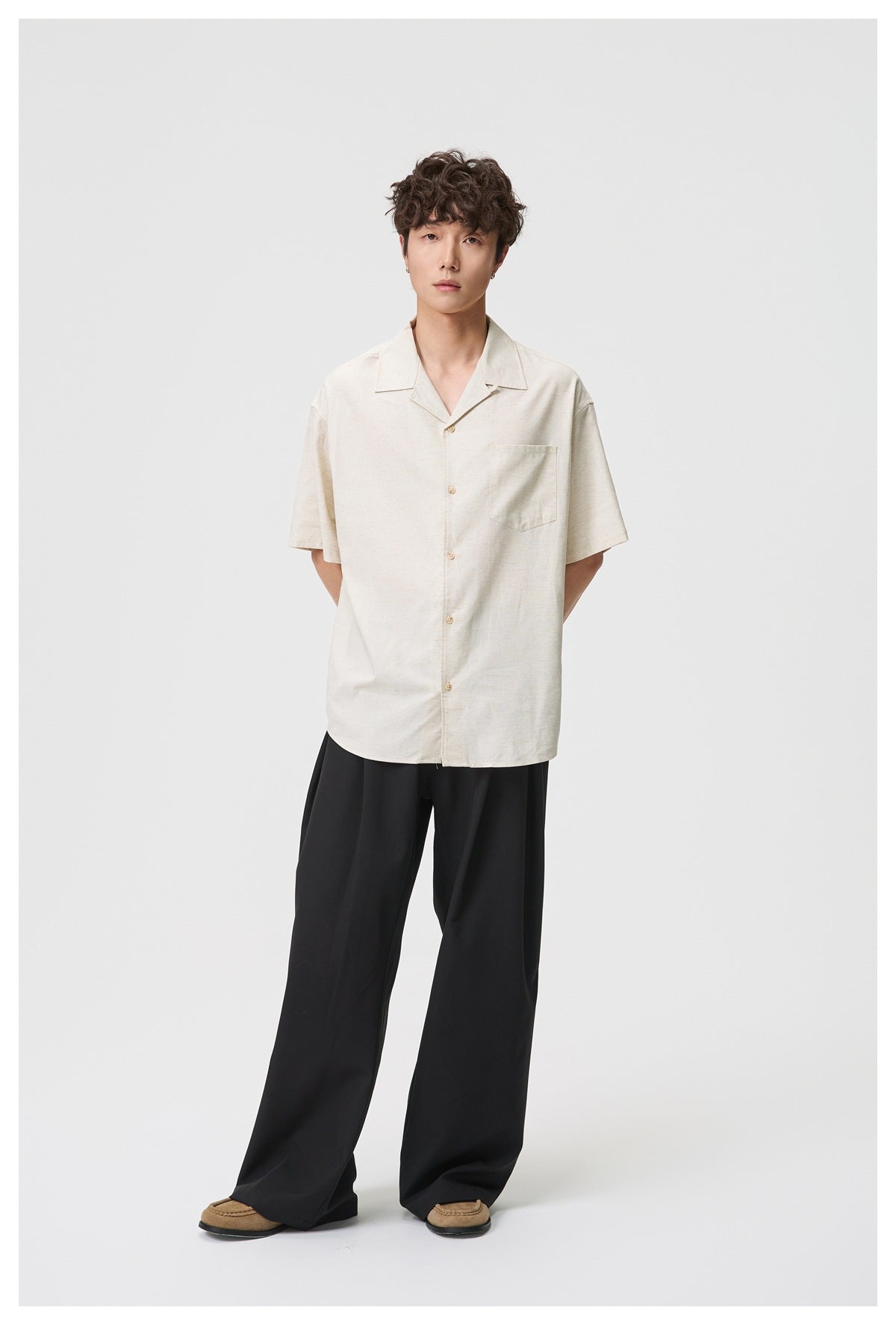 Full-length view of a male model wearing an off-white short-sleeve button-up shirt and loose black trousers.