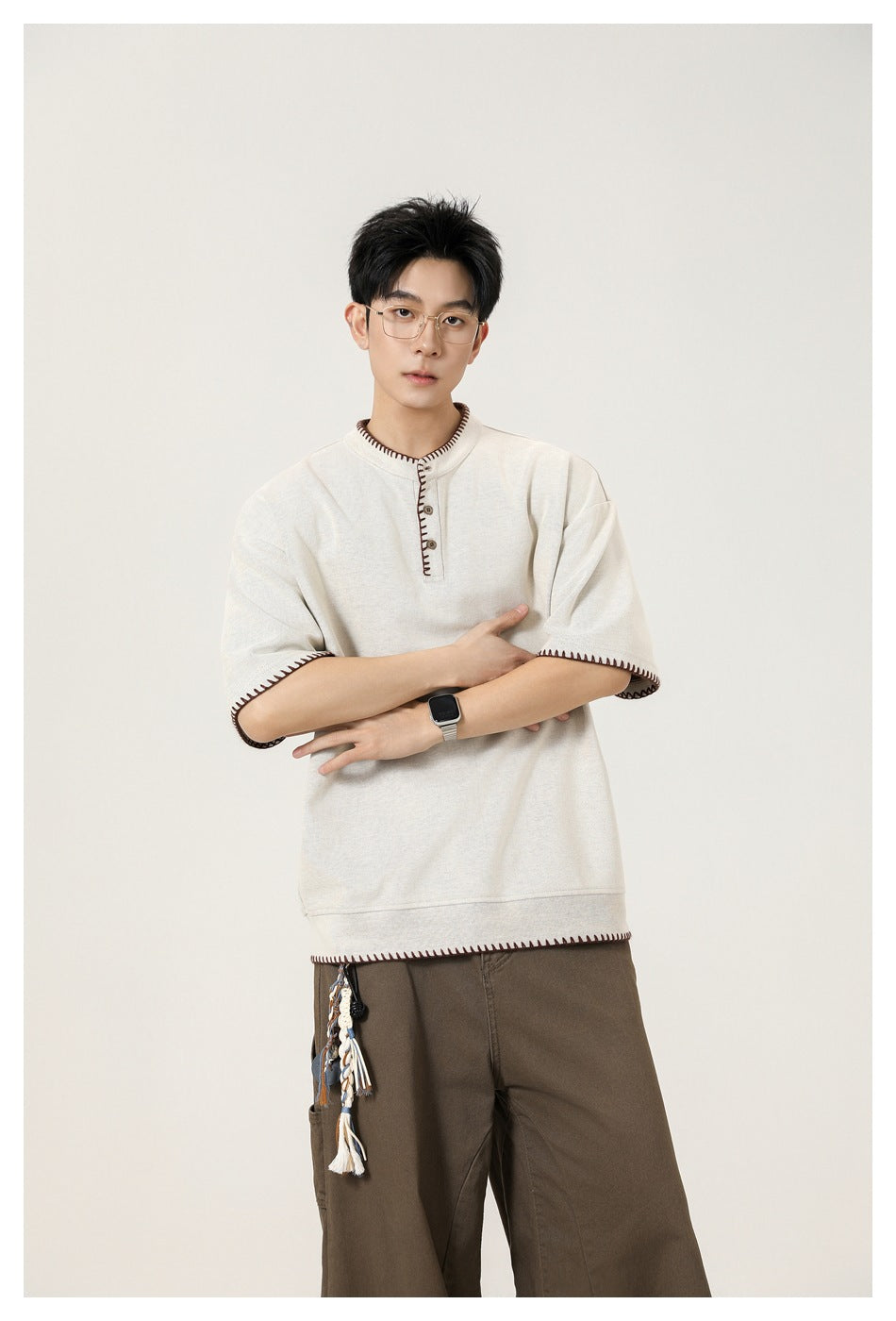 Model wearing an ivory henley with dark stitched trim, arms crossed, and brown trousers.