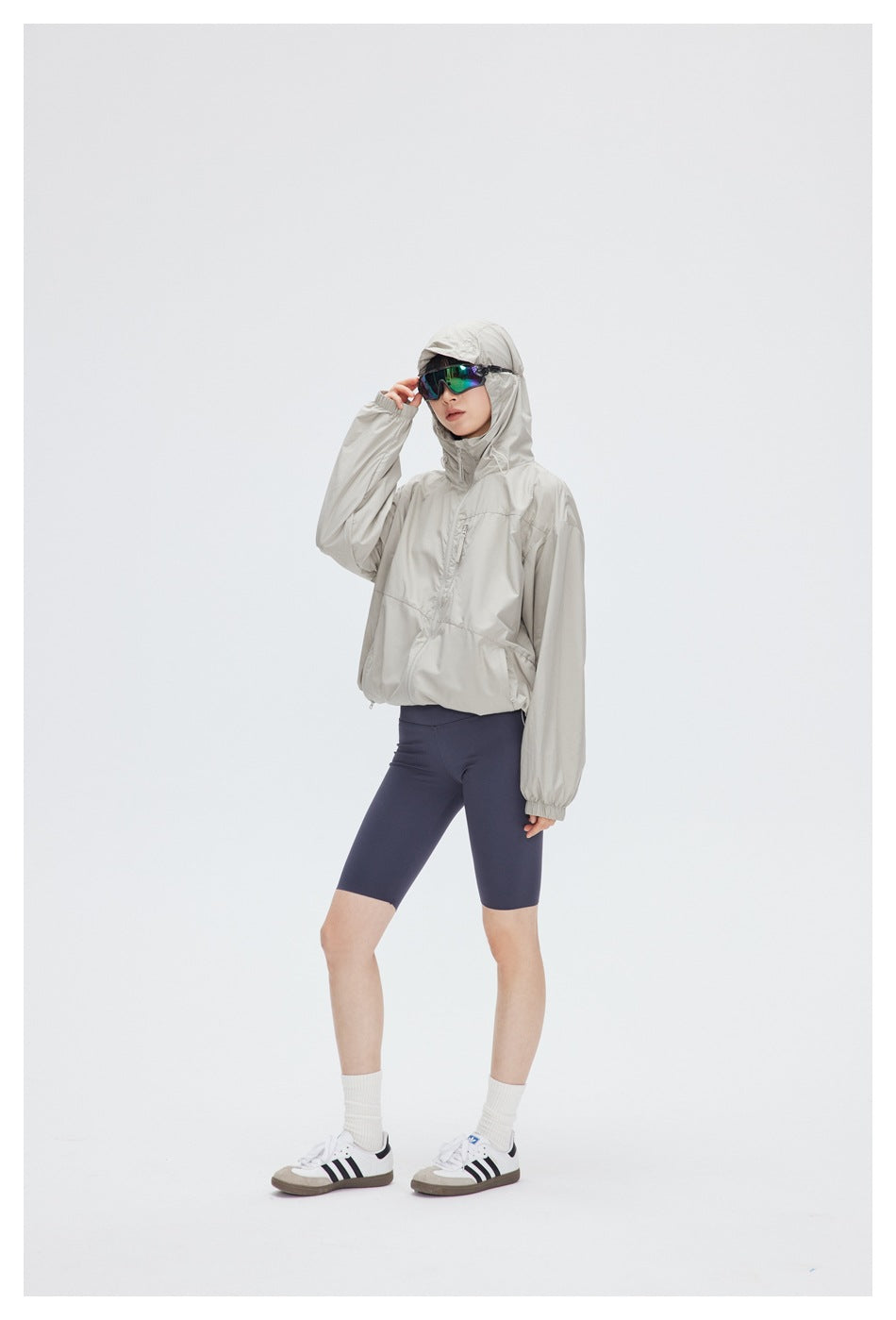 Model wearing a light beige hooded windbreaker and navy cycling shorts, shown full length.