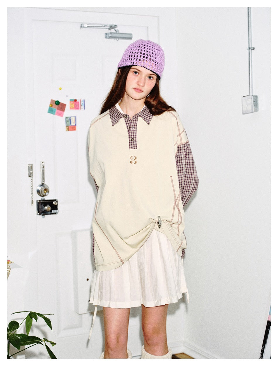 Model wearing a cream oversized top with brown plaid collar and sleeves, gathered at the side over a white pleated skirt.