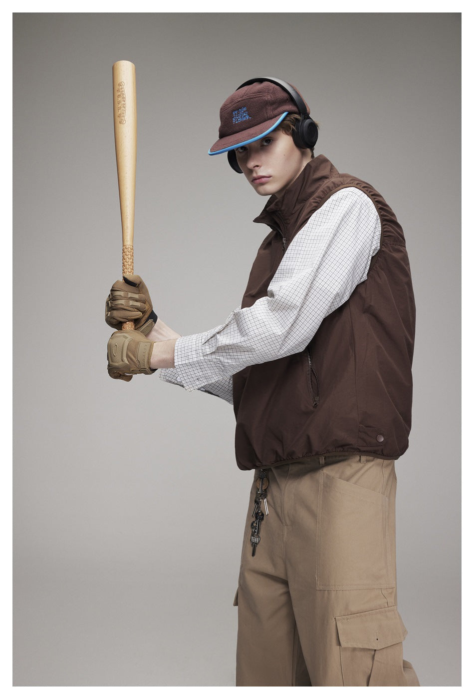 Model wearing a brown sleeveless zip vest over a checked shirt with khaki cargo pants, cap, and headphones.