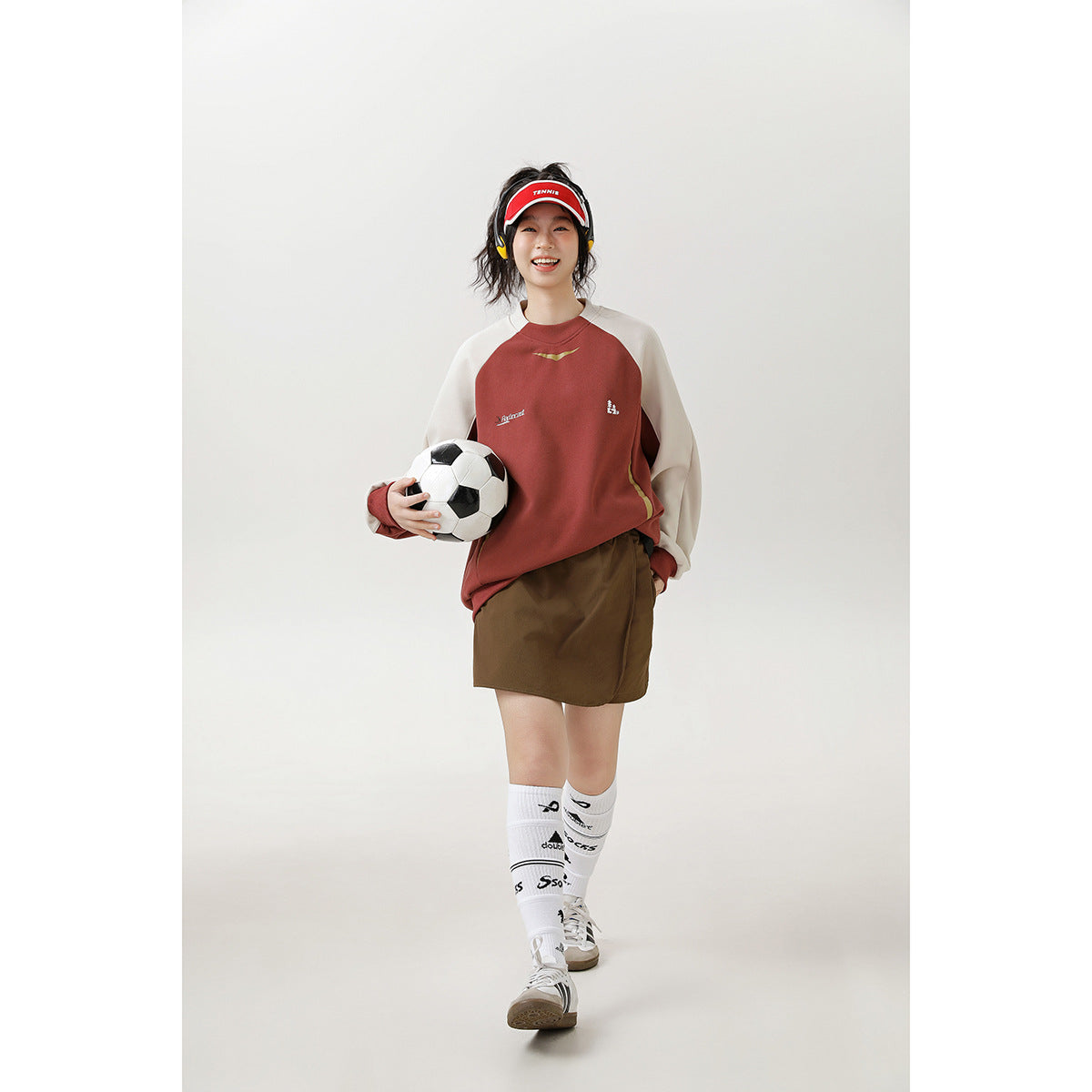 Full-length view of a woman in a rust-red and cream sweatshirt, brown skirt, and white knee socks holding a soccer ball.