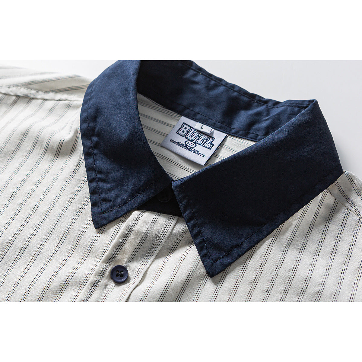 Close-up of a cream shirt with fine dark stripes, navy pointed collar, button, and sewn neck label.