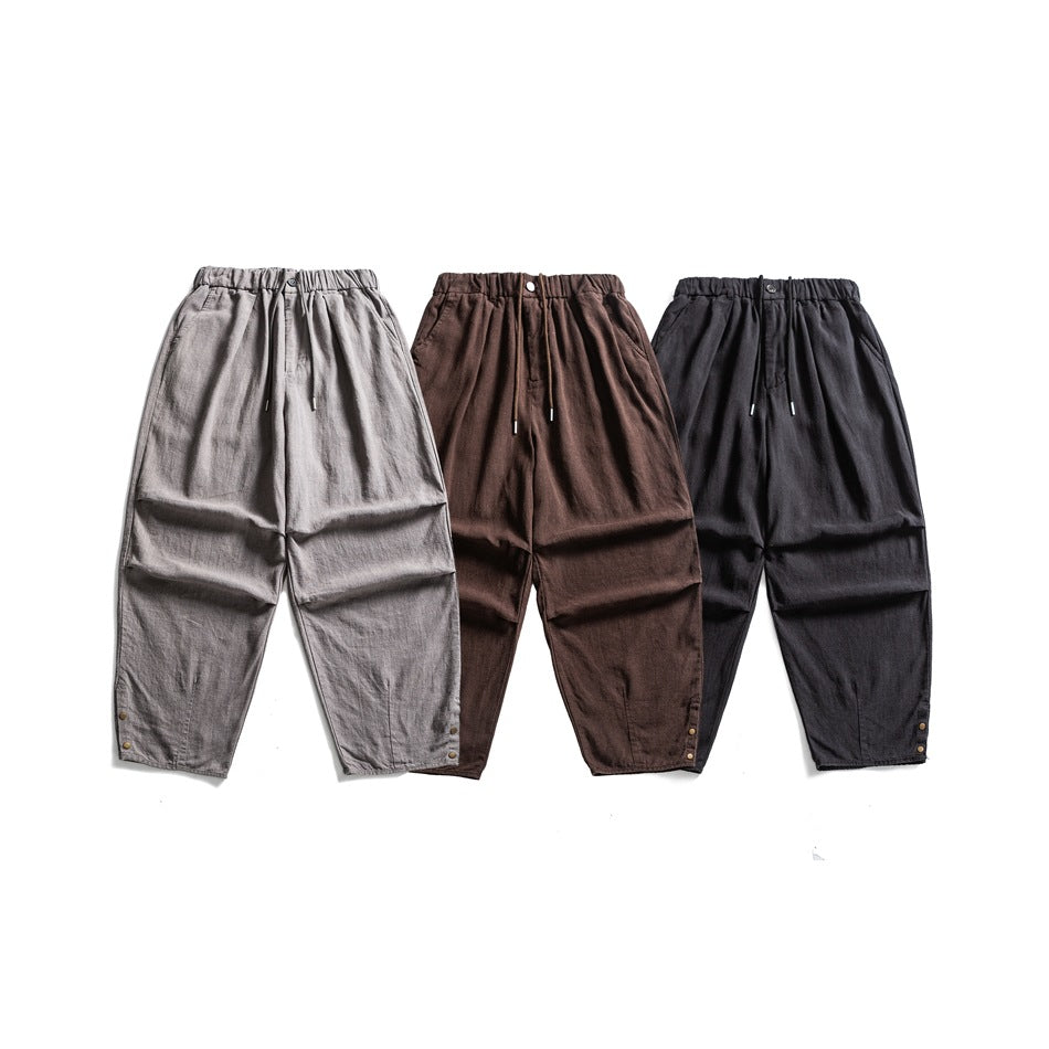 Front view of three relaxed trousers in light gray, brown, and black, with elastic waists and ankle snaps.
