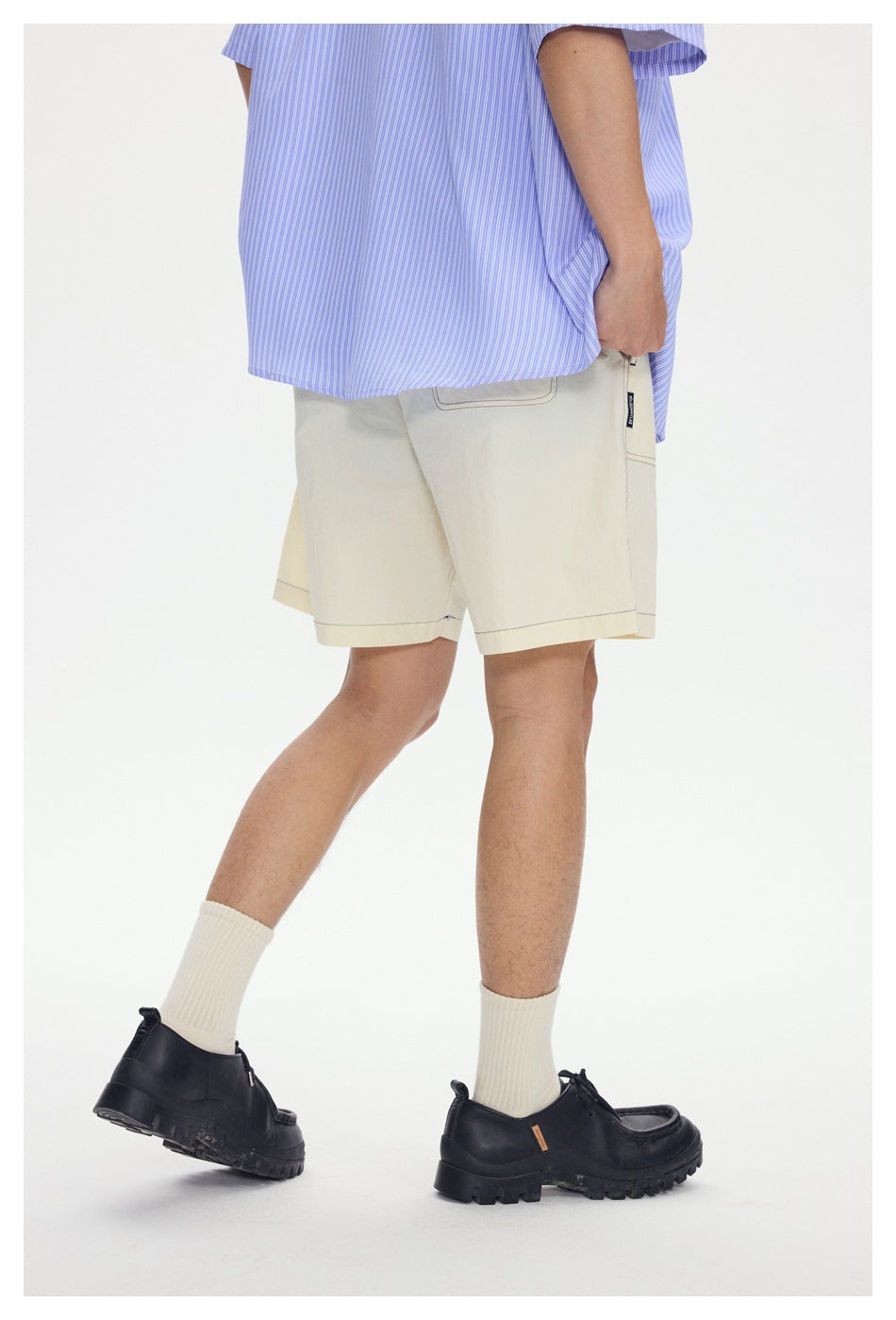 Side and rear view of a model wearing cream knee-length shorts with stitched pockets and a blue striped shirt.