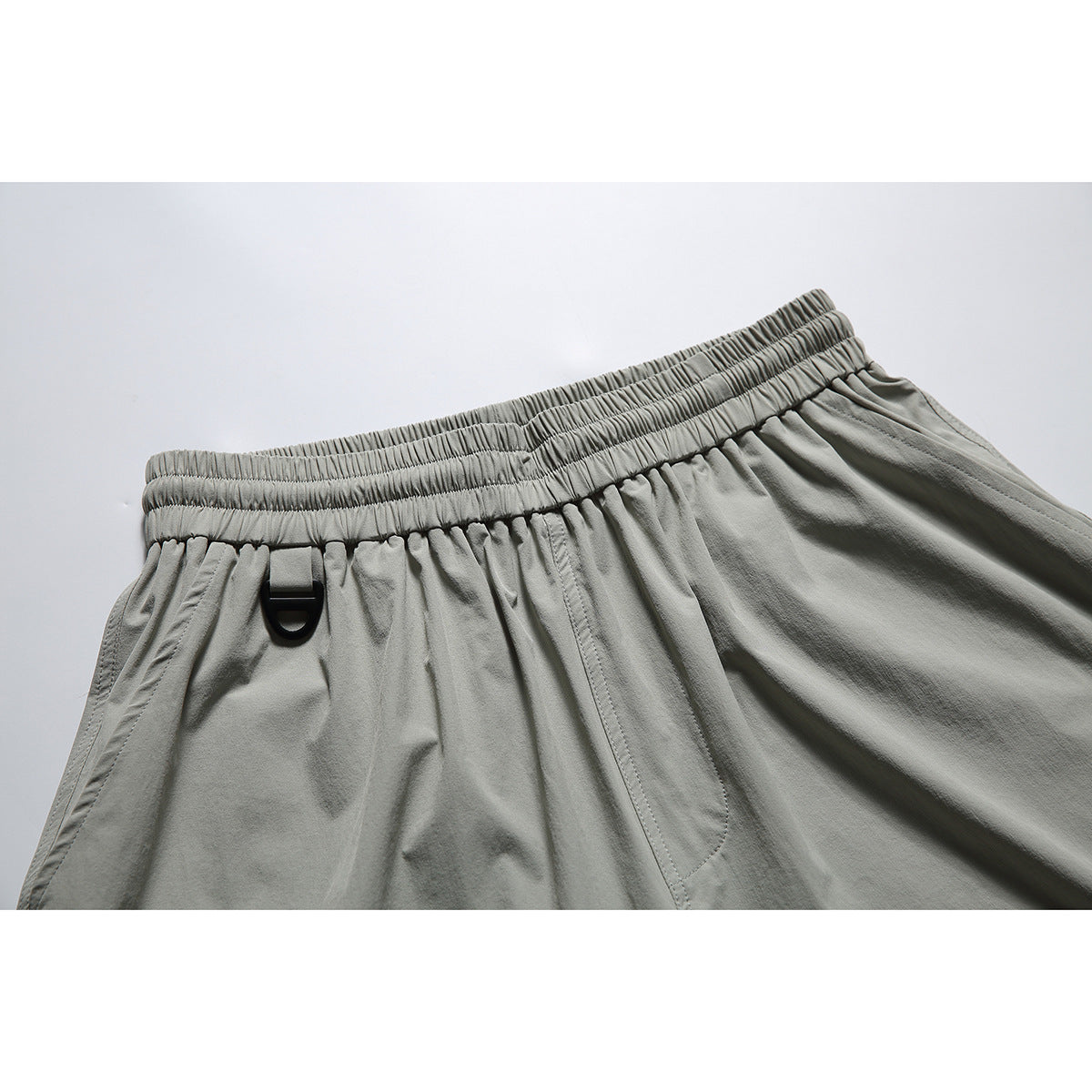 Close-up of gray-green shorts showing the gathered elastic waistband, black D-ring, and front pocket seam.