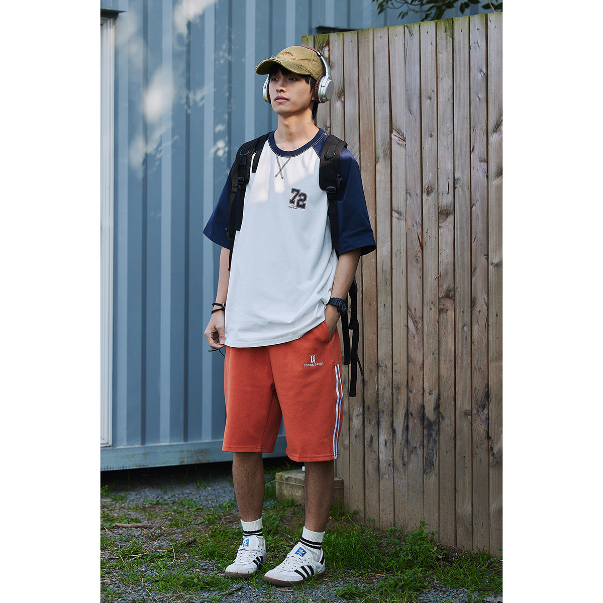 Full-length model wearing a white and navy raglan T-shirt with a chest number graphic and rust-orange shorts.