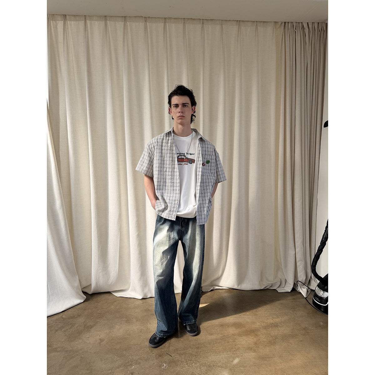 Full-length front view of a man wearing a blue-gray plaid short-sleeve shirt over a white graphic tee and loose dark jeans.