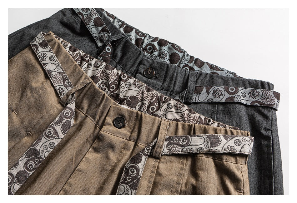 Two pairs of trousers in charcoal and tan, shown overlapping with patterned trim, belt loops, and button closures.