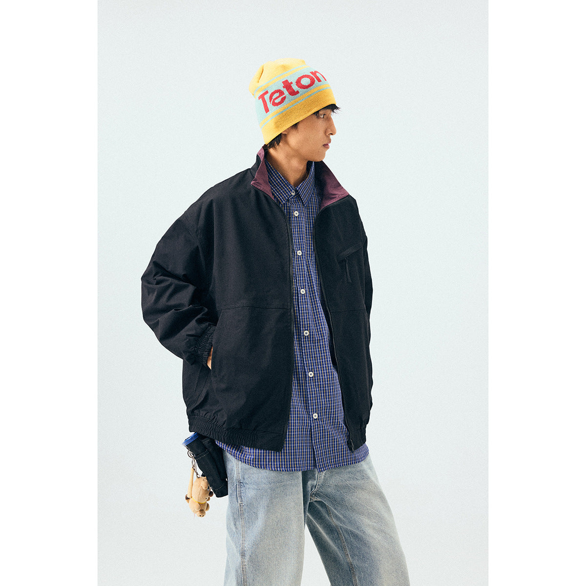 Model wearing a black zip-front windbreaker with a plum collar, blue checked shirt, and light jeans.