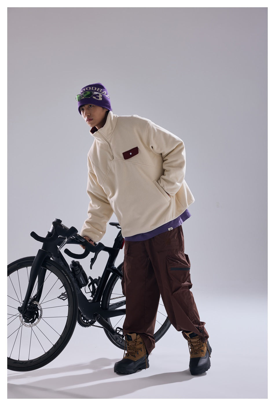 Model wearing an ivory quarter-zip pullover and brown cargo pants while leaning on a black bicycle.