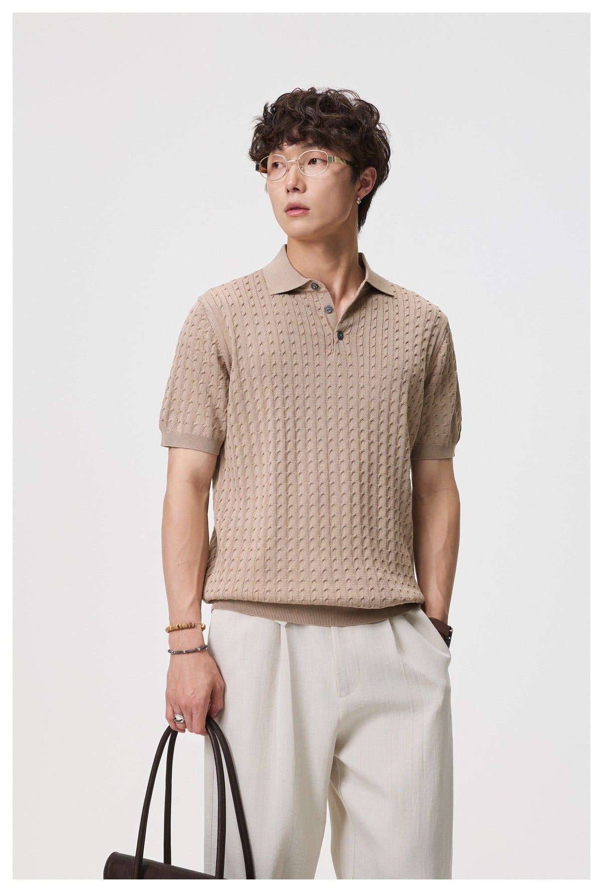 Man wearing camel cable-knit short-sleeve polo with cream trousers, holding a brown handbag, front view