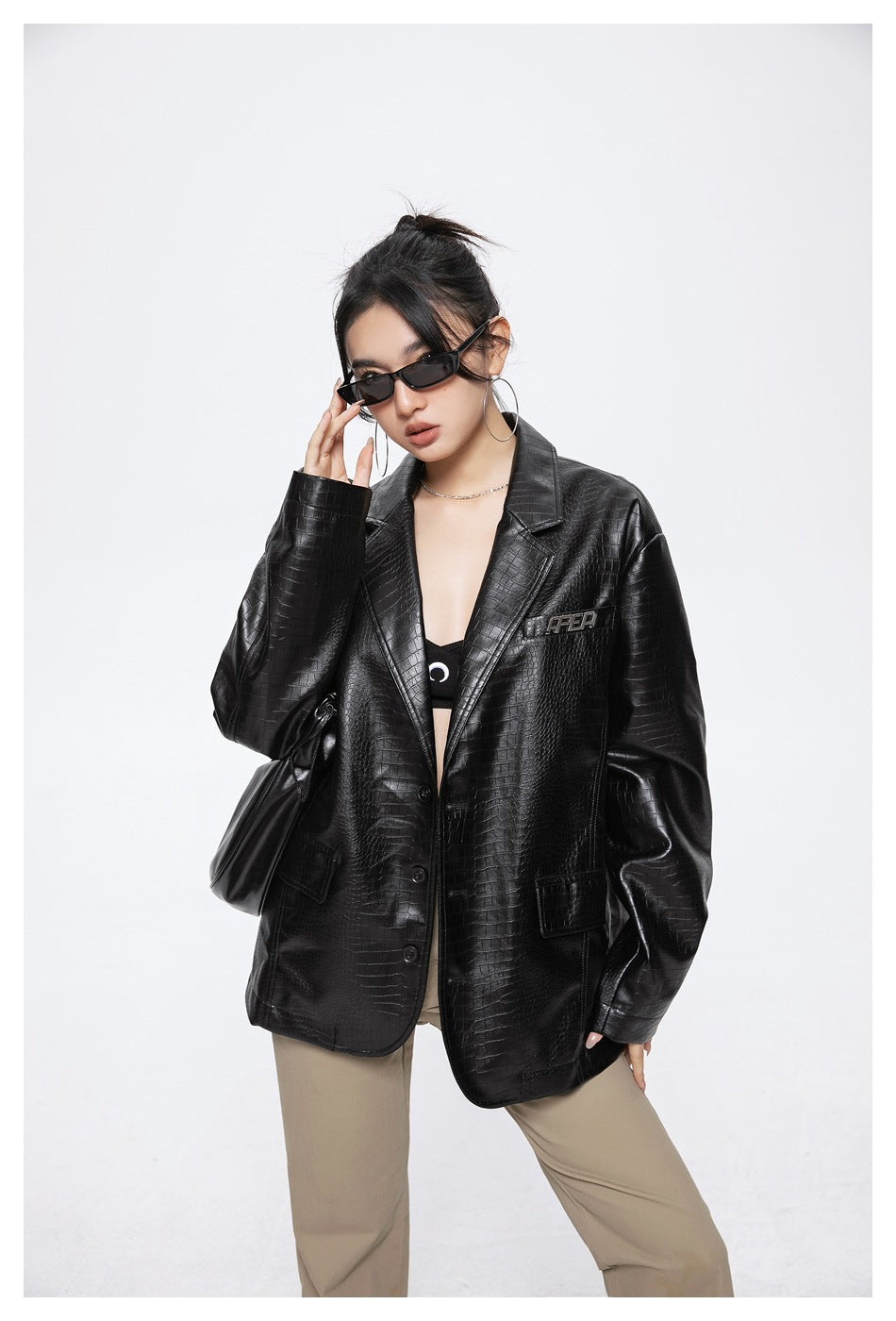 Model wearing the black crocodile-embossed jacket open over a dark top with khaki pants, hand at sunglasses