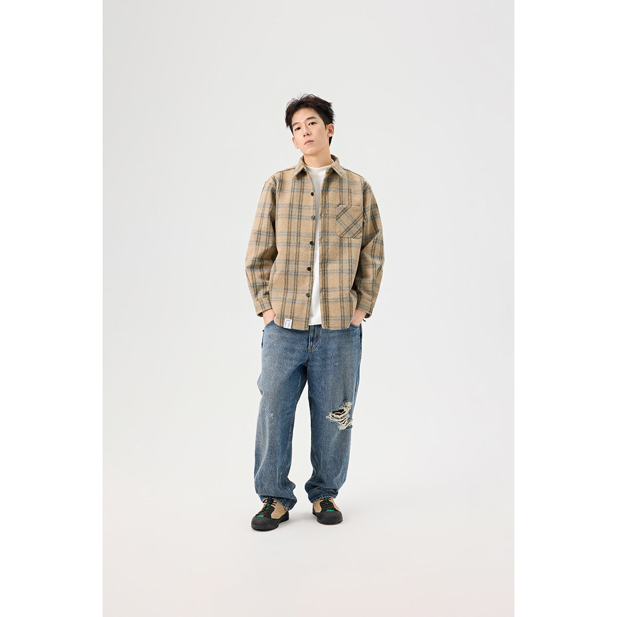 A man models a tan plaid overshirt, white tee, and loose blue jeans, shown nearly full length.