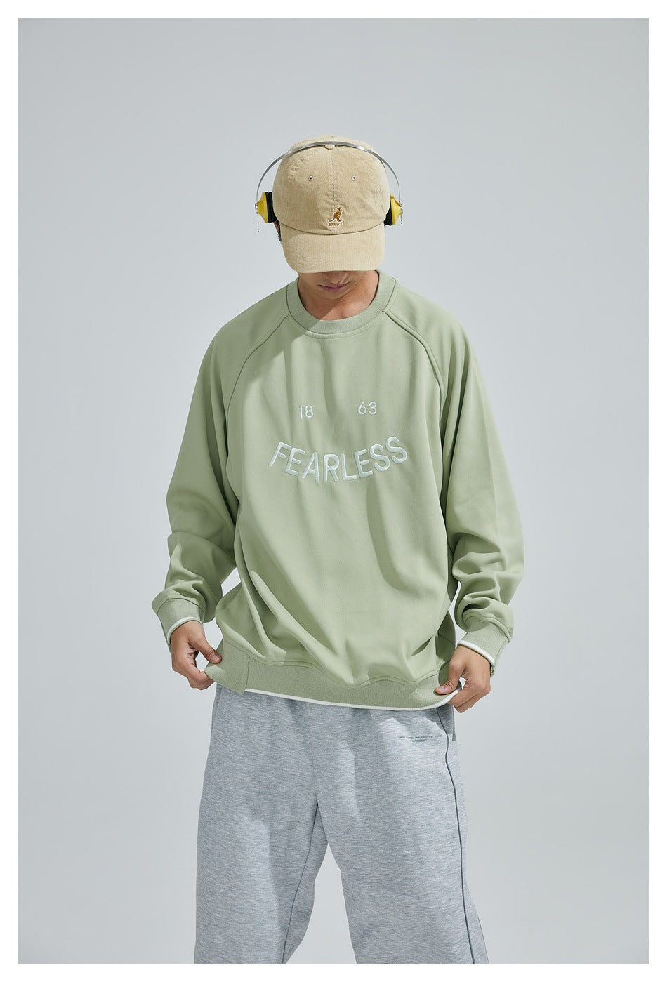 Model looking down while wearing a sage-green sweatshirt with white “18 63 FEARLESS” embroidery.