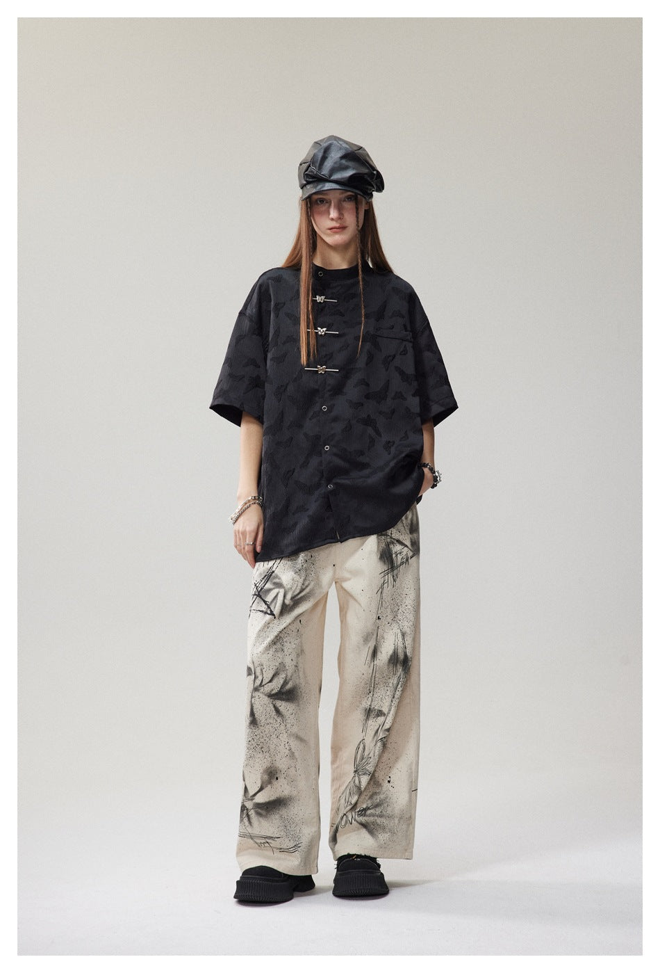 Model wearing the black butterfly-pattern short-sleeve shirt with metal toggle closures, a black cap and painted cream pants