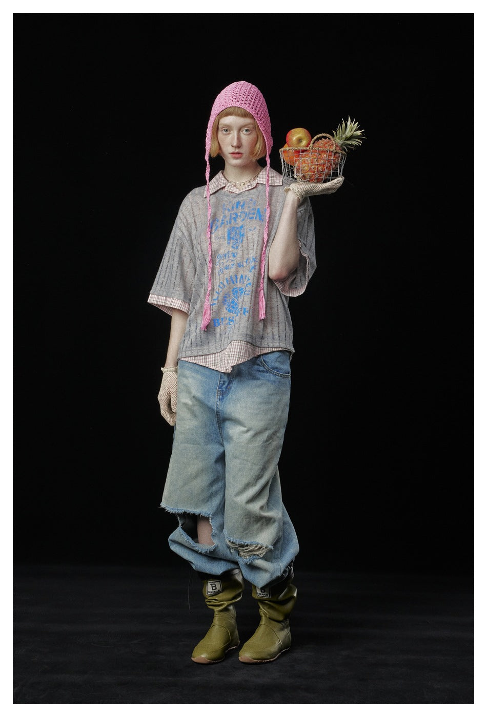 Model wearing a loose gray graphic T-shirt over a pink checked shirt, with wide distressed jeans and a pink knit bonnet.