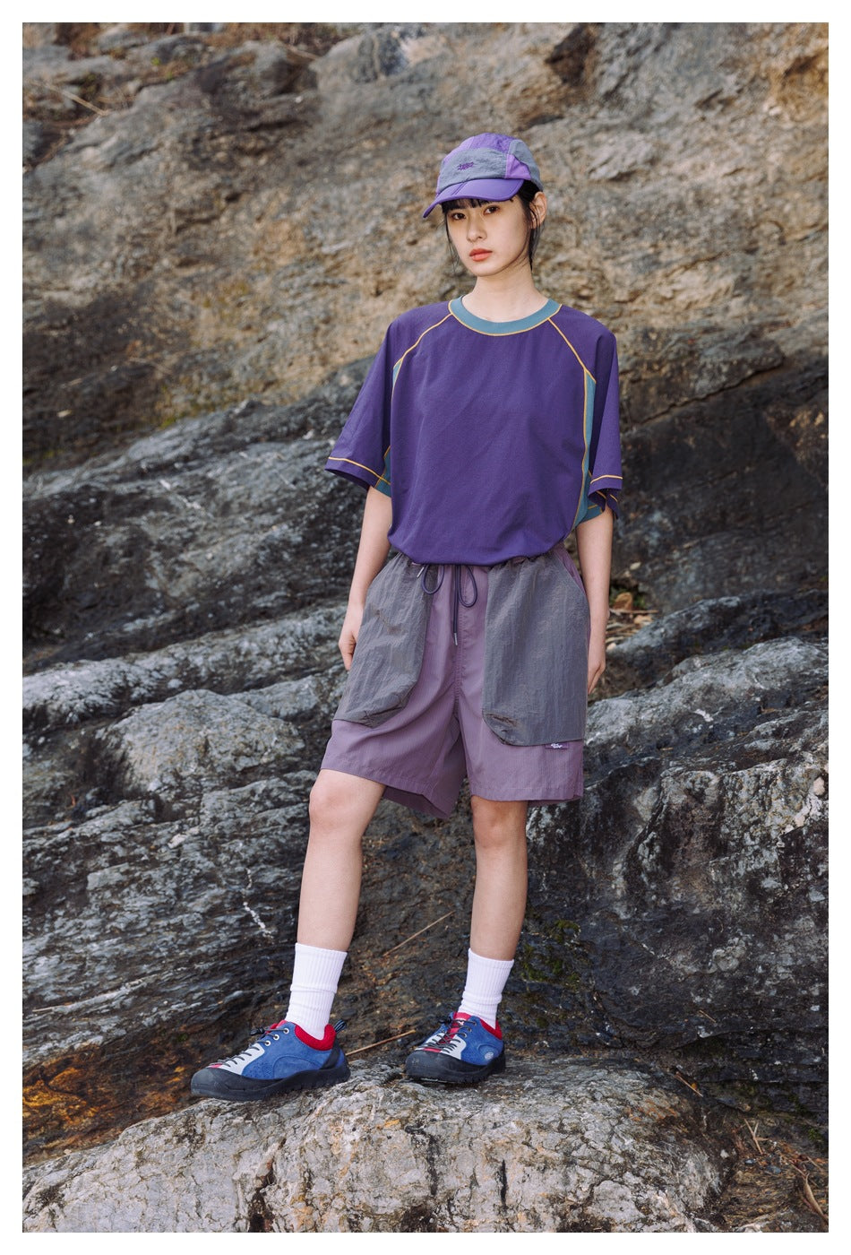Front view of a woman wearing a purple color-block T-shirt, lilac utility shorts and a matching cap outdoors on rocks.